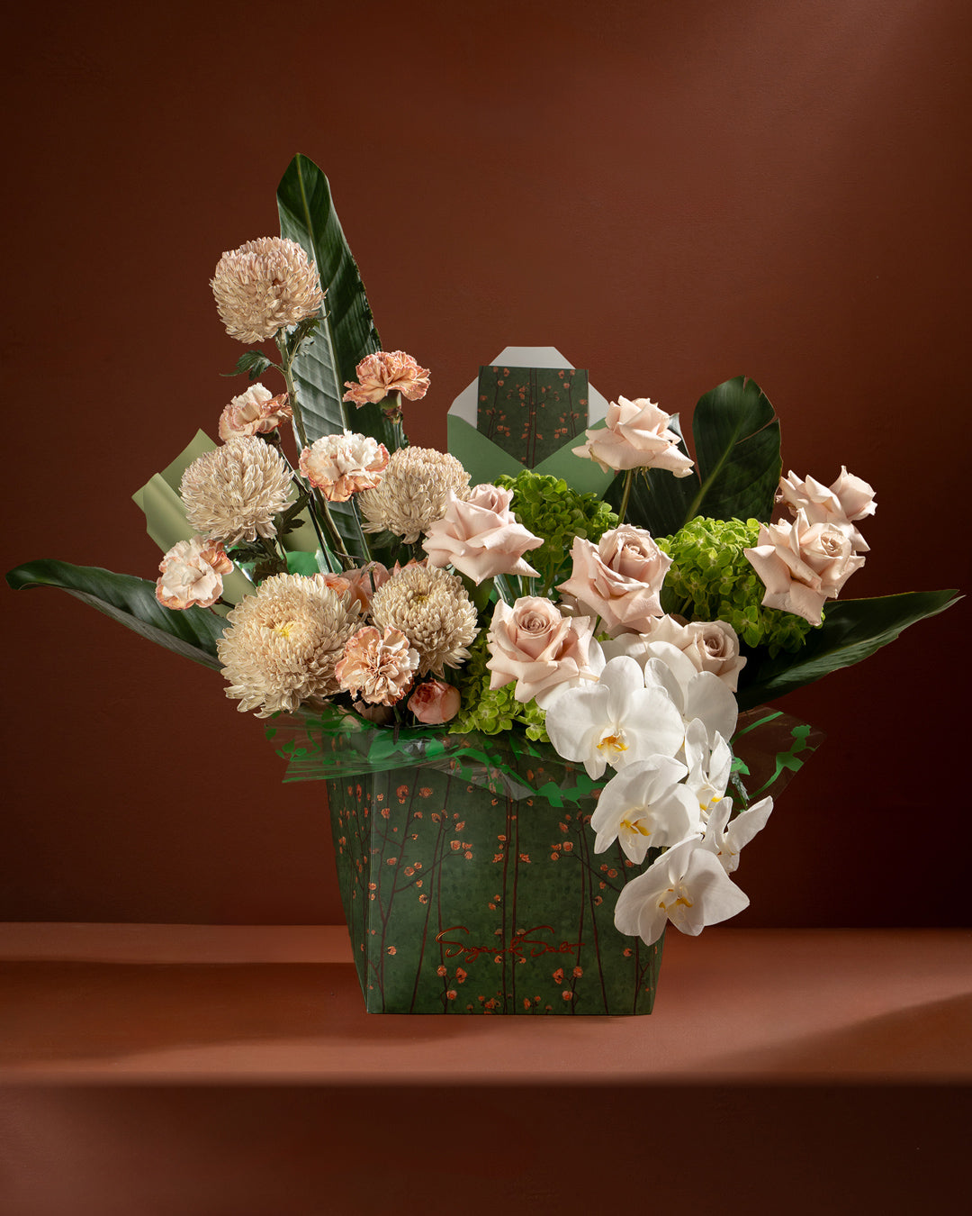 Floral arrangement with white and pink flowers in a green box against a brown background